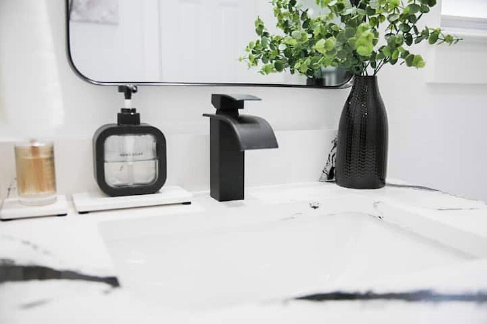 Une salle de bain avec un lavabo blanc, un distributeur de savon noir, une bouteille ronde et une plante en pot.