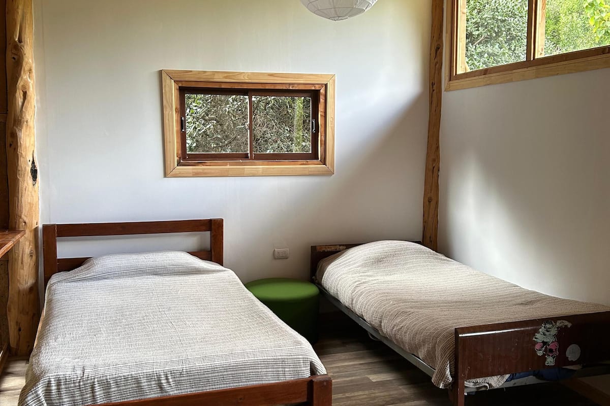 Une pièce avec deux lits en bois, un pouf vert, une fenêtre donnant sur un paysage verdoyant et un luminaire suspendu.