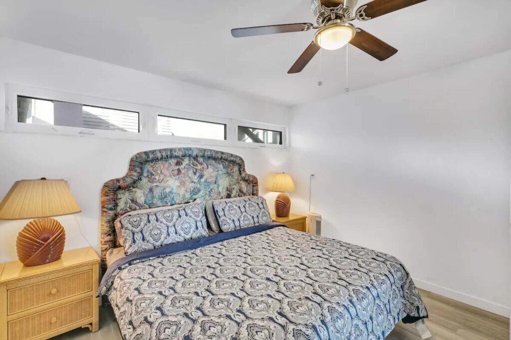 A bedroom with a bed, a ceiling fan, a nightstand with a lamp, and a wall-mounted shelf.