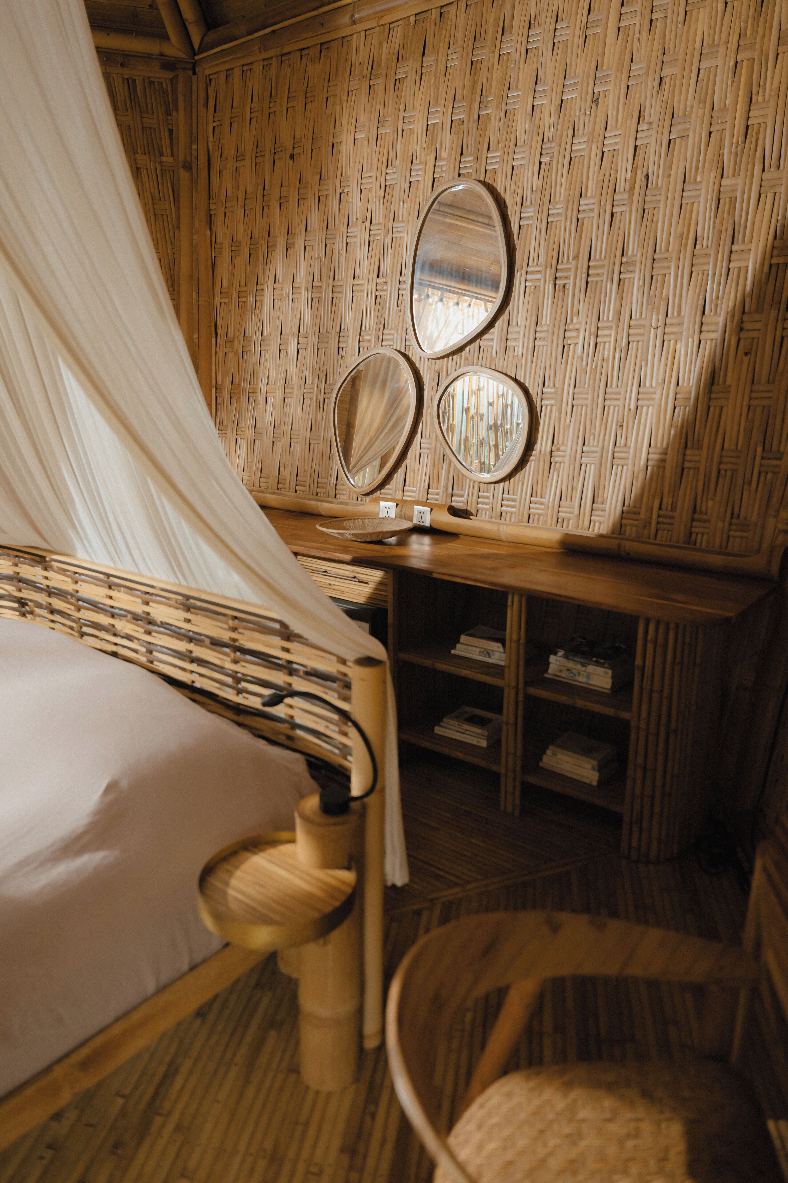 A room with a bed, a chair, a bedside table with a mirror, and a shelf with books.