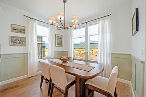 A dining room with a wooden table, four chairs, a chandelier, and large windows with a view of a landscape.