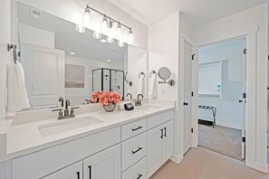 A modern bathroom with a large mirror, white vanity, and a shower area visible through an open door.