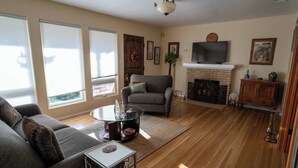Sala de estar com sofá, poltrona, mesa de centro de vidro, lareira, televisão e piso de madeira.