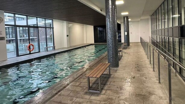 An indoor swimming pool with clear water, wooden benches, and large windows.