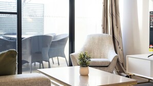 A modern hotel room with a large window, a sofa, a chair, a small table with a plant, and a TV stand.