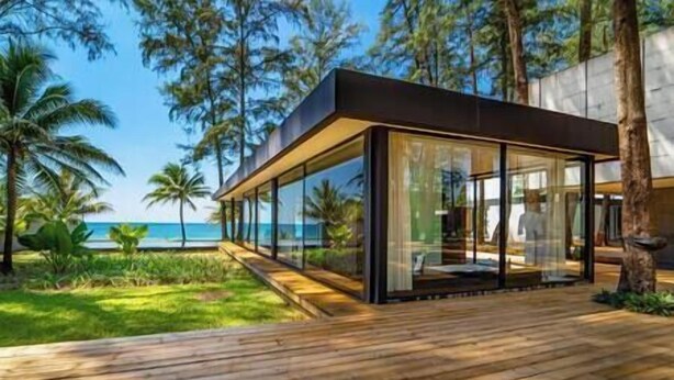 Una casa moderna de vidrio con terraza de madera, rodeada de vegetación tropical y con vistas a una playa.