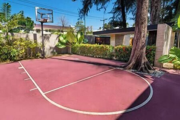 An outdoor basketball court with a red surface, surrounded by greenery and a building in the background.