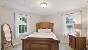 A bedroom with a wooden bed, a dresser, a mirror, and two windows with curtains.