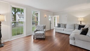 A spacious living room with hardwood flooring, a patterned armchair, a grey sofa, and large windows offering a view of the outside.