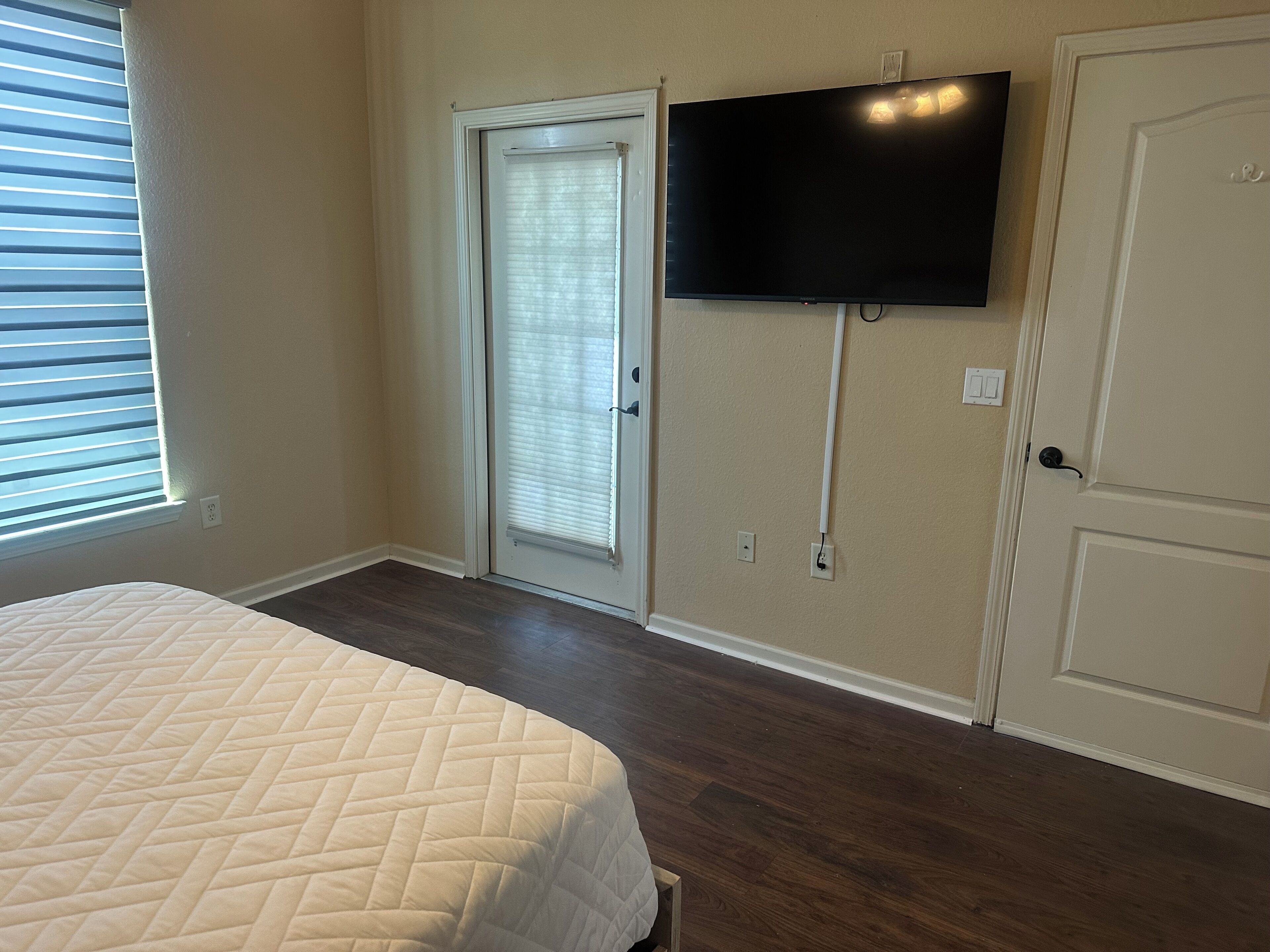 Un dormitorio con cama, una puerta corrediza de vidrio, un televisor montado en la pared y una puerta con picaporte.
