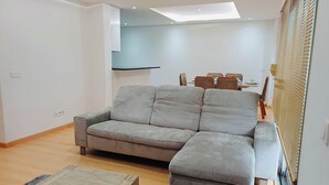 Een moderne woonkamer met een grijze modulaire bank, een eettafel met stoelen en een groot raam met zonwering.