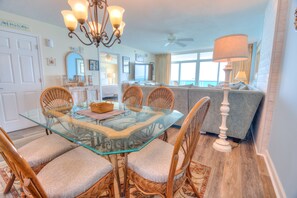 A dining area with a glass table, wicker chairs, a chandelier, and a sofa in the background.