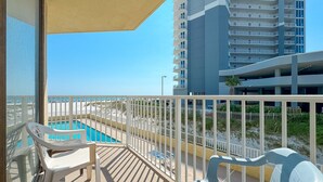 Un balcón con vistas a una piscina y una playa, sillas de plástico blanco y un edificio alto al fondo.