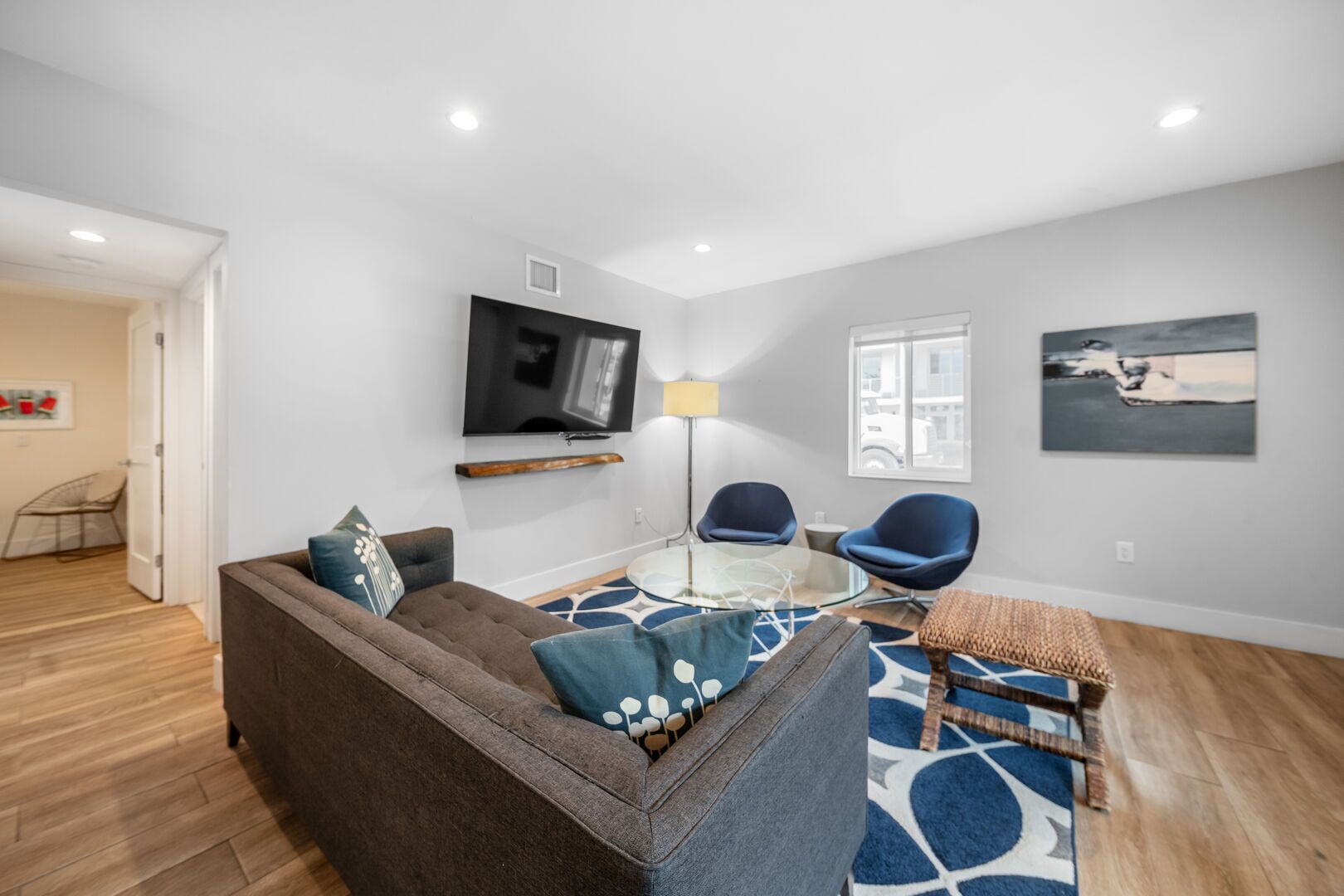 Een moderne woonkamer met een bruine bank, twee blauwe fauteuils, een glazen salontafel, een aan de muur bevestigde televisie en een kamerkleed met dessin.