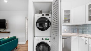 A laundry room with two stacked front-loading washing machines, a teal sofa, a flat-screen TV, and a kitchen area with white cabinets and blue-tiled backsplash.