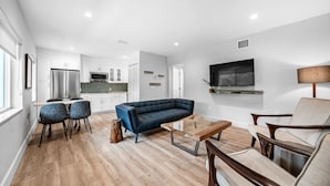 Una sala moderna con un sofá azul, una mesa de centro de madera y dos sillones. Se ve una cocina con armarios blancos y un refrigerador de acero inoxidable al fondo.