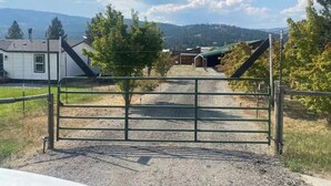 A gated rural property with a gravel driveway, a white building, and a barn in the background. - Charming 2-bedroom house in beautiful Republic with infrared sauna. (Republic)