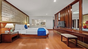 A hotel room with a large bed, a wooden headboard, a bedside table with a lamp, a bench, and a large mirror. - Sandals Stay Hotel (Da Nang)