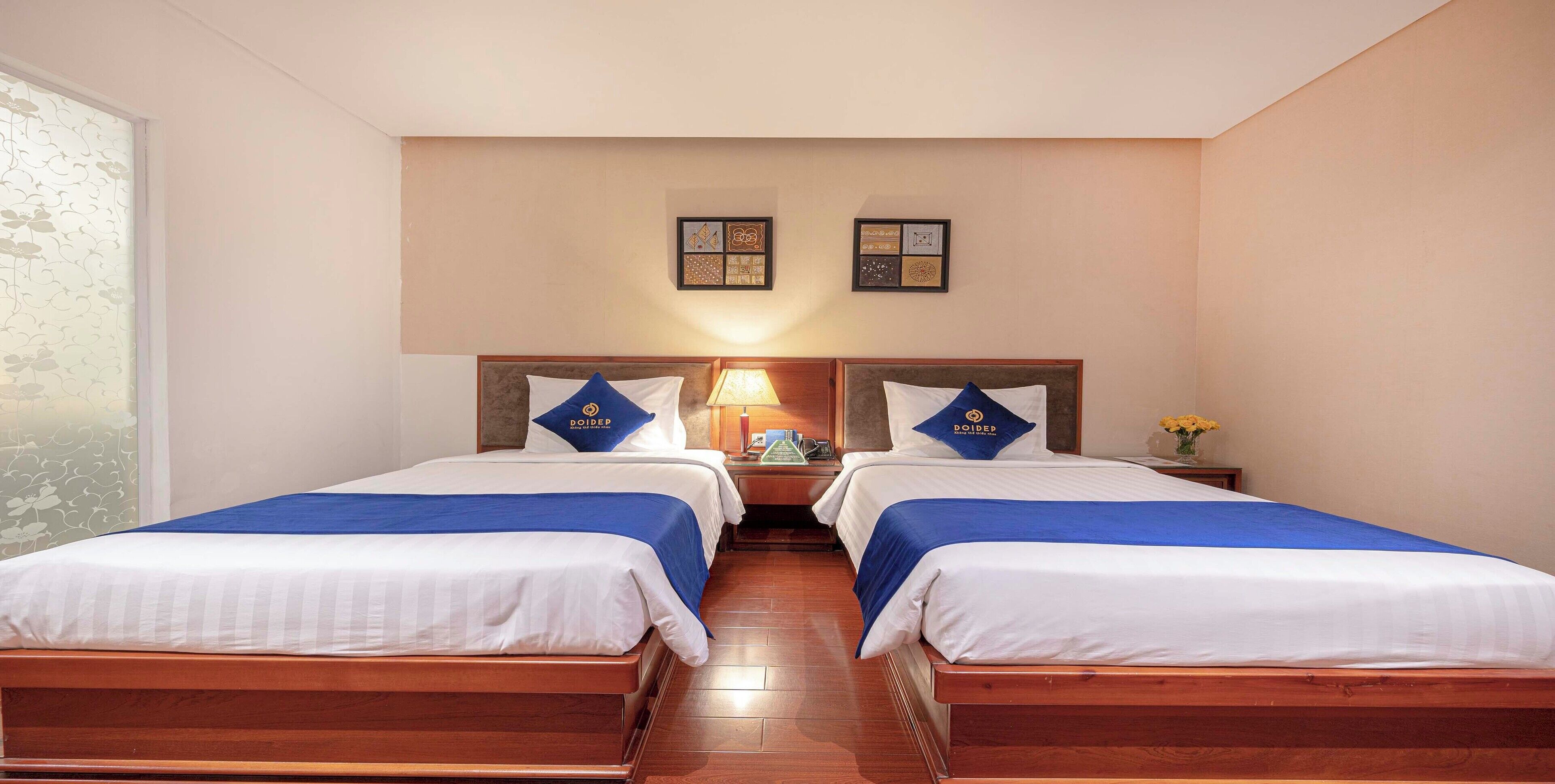 A hotel room with two beds, each with a blue bedspread and a pillow with the hotel logo. There are framed pictures on the wall above the beds and a nightstand with a lamp between them.