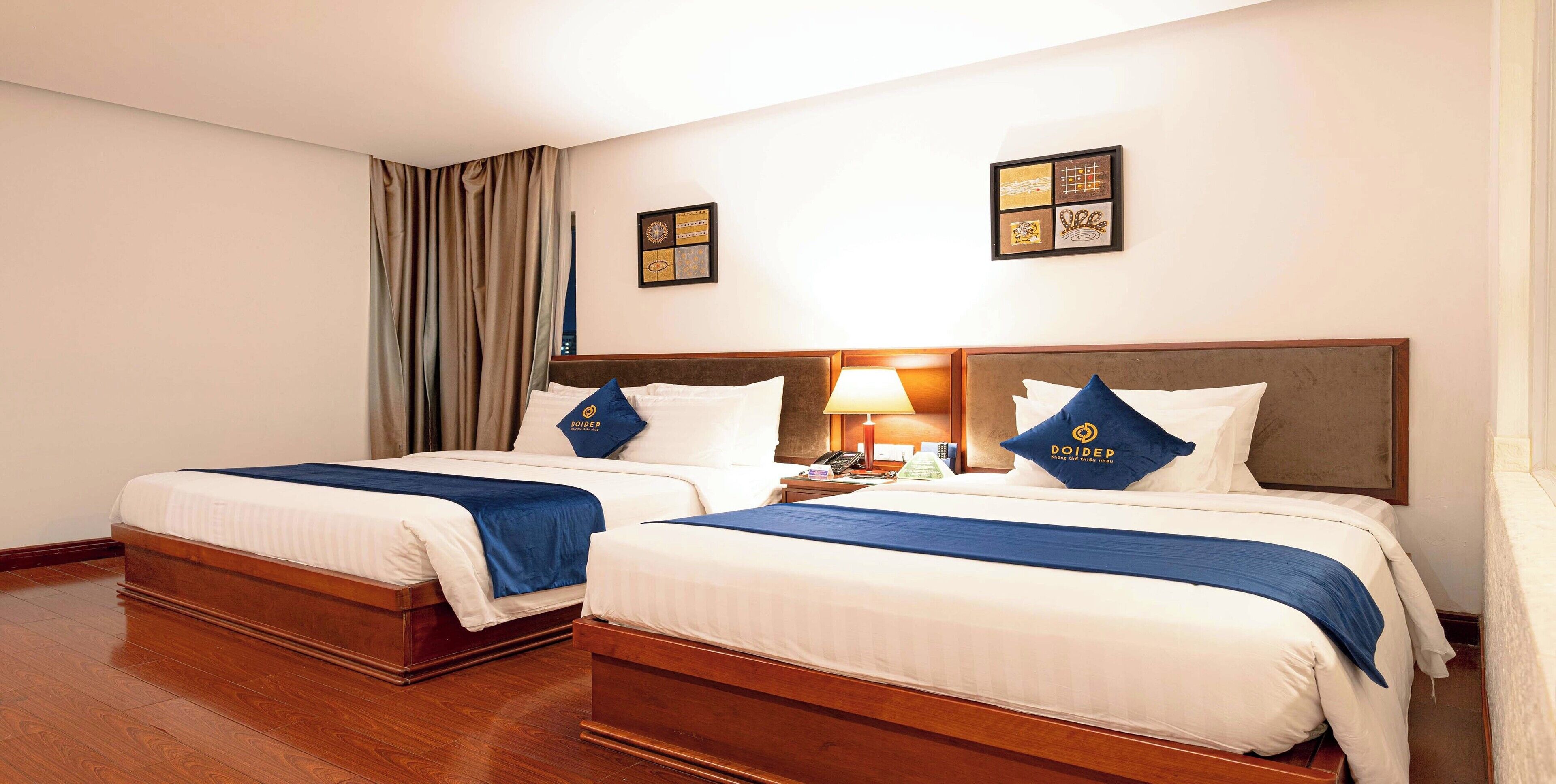 A hotel room with two beds, each with a blue bedspread and matching pillows, a wooden headboard, a nightstand with a lamp, and a wall-mounted artwork.