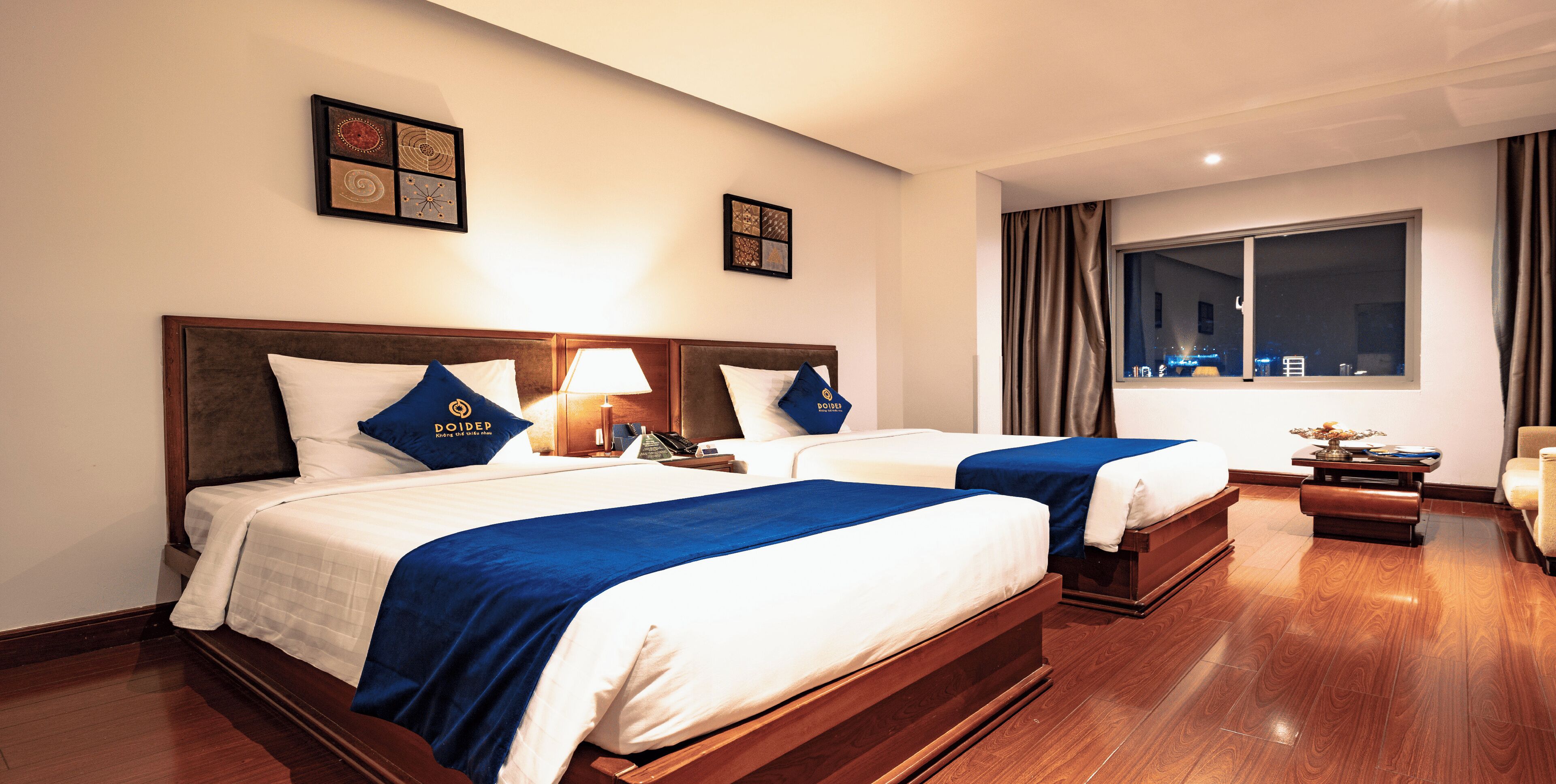 A hotel room with two beds, each with a blue bedspread, white linens, and a pillow with the hotel logo. There is a nightstand with a lamp, a window with curtains, and a view of the cityscape at night.