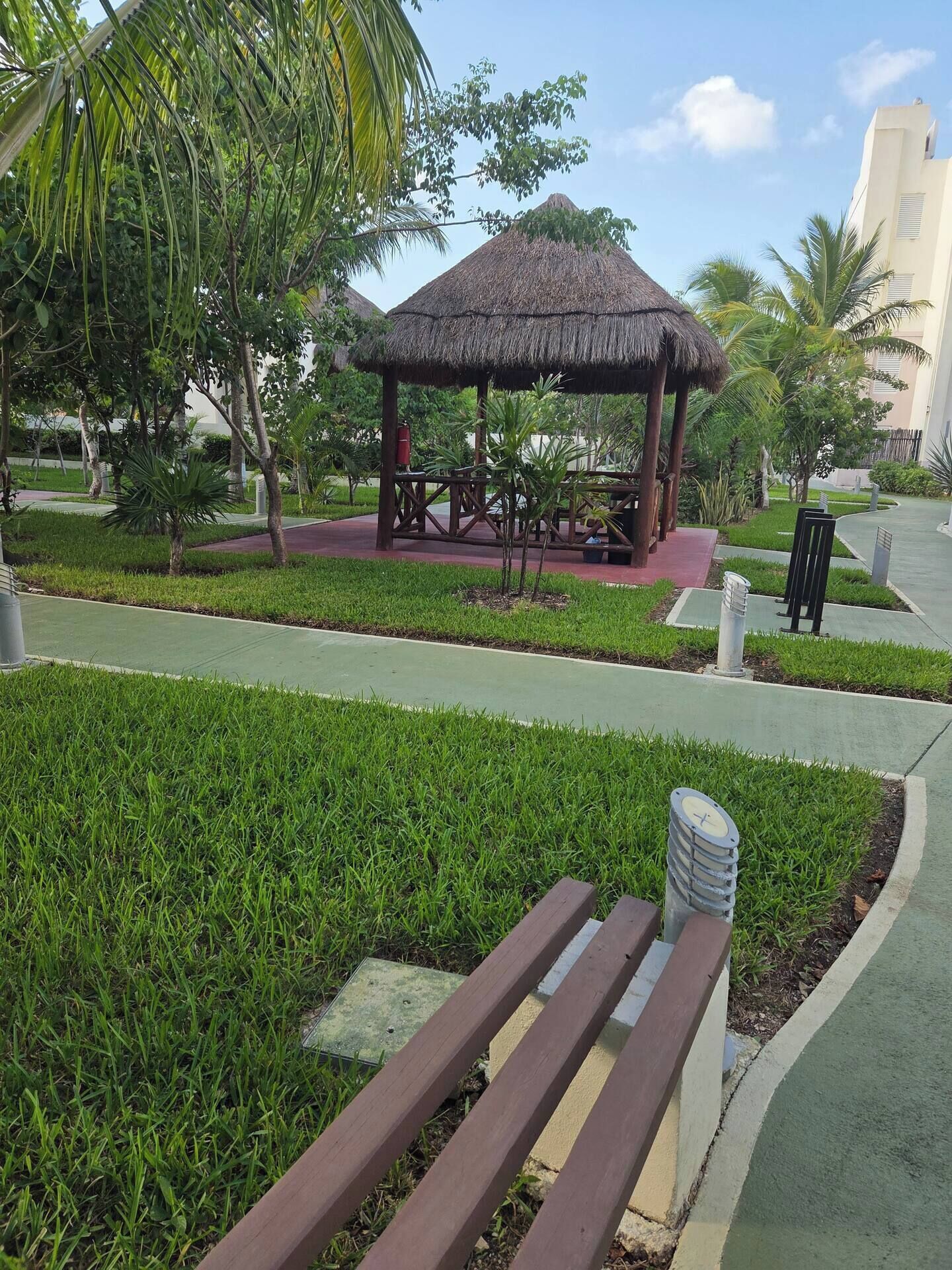 A park area with a thatched-roof gazebo, a paved walkway, and green grass.