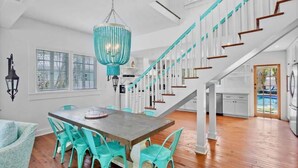 Un vaste espace dédié à la salle à manger, avec une table en bois et des chaises turquoise, un escalier agrémenté de touches de couleur vert émeraude, et un grand lustre.