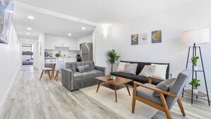A modern living room with a grey sofa, a wooden coffee table, and a grey armchair. There is a kitchen in the background with white cabinets and a refrigerator.