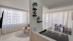 Una cocina moderna con una isla central, estantes empotrados con plantas, un comedor con mesa y sillas, y ventanas amplias con cortinas traslúcidas.