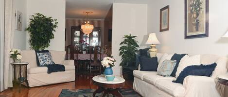 A living room with two sofas, a coffee table, a chandelier, and a dining area in the background.