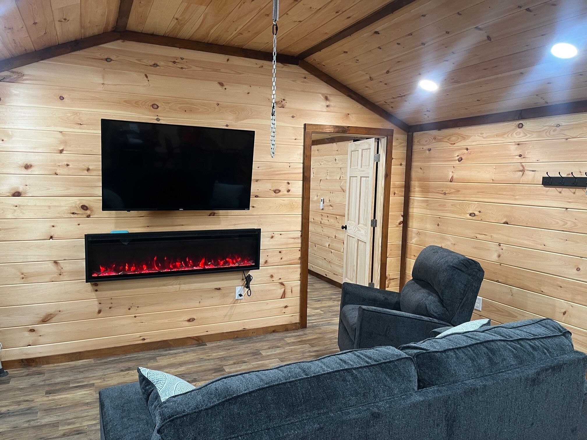 A cozy room with a wooden interior, a flat-screen TV mounted on the wall, a fireplace below it, and a grey sofa.