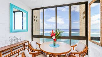 A round table with wicker chairs and a wooden bench, a vase with flowers, and a view of the ocean through large windows.