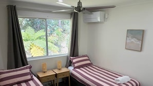 Une chambre avec deux lits simples, un bureau, une chaise, un ventilateur de plafond et une fenêtre donnant sur un paysage verdoyant.