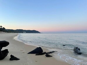 Ein Strand mit großen Felsen im Vordergrund, einem Sandufer und ruhigem Wasser.