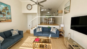 Una sala de estar con un sofá, una mesa de centro y un televisor. Se ve una escalera que conduce a un nivel superior con un comedor.