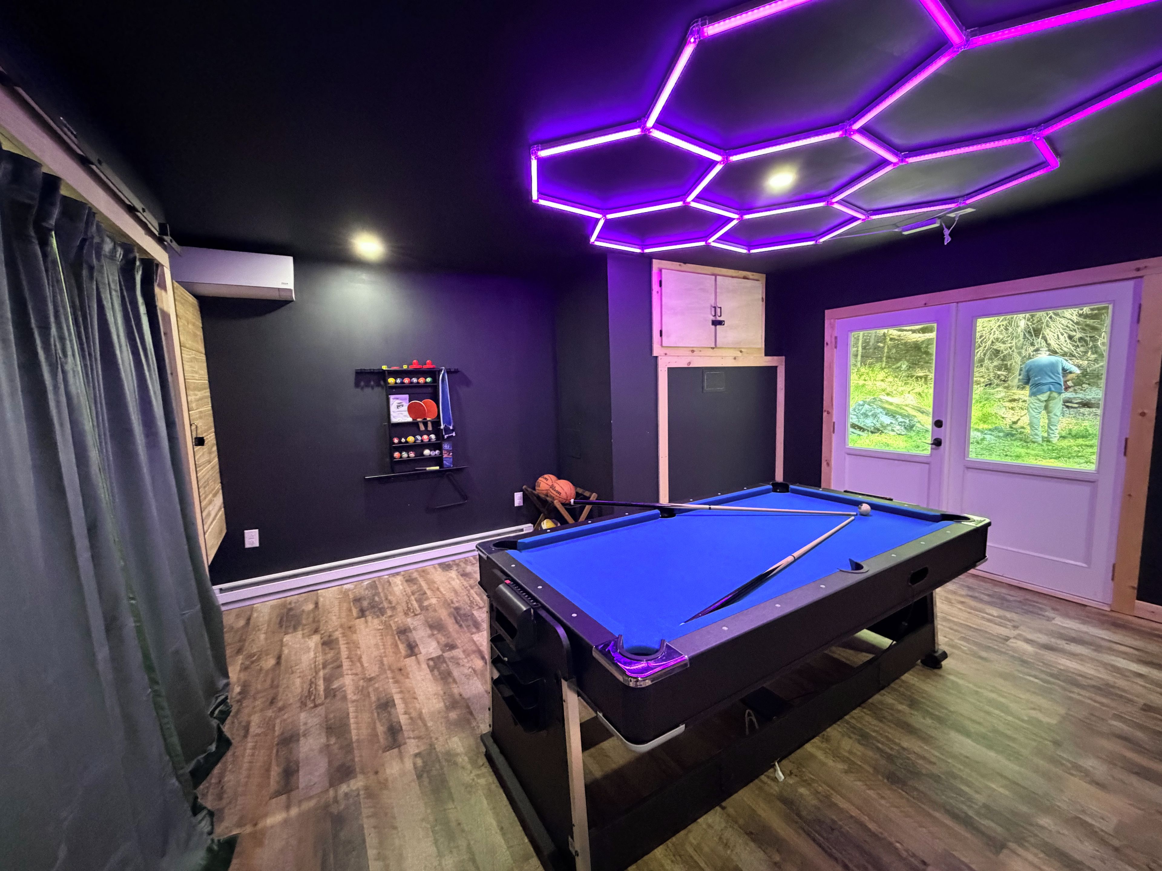 A pool table in a room with a wooden floor, a door with a window, and a wall-mounted rack with billiard balls.