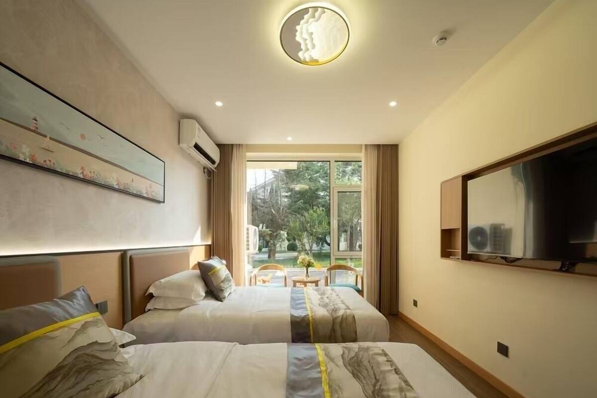 A hotel room with two beds, a large window, a flat-screen TV, and a ceiling light fixture.