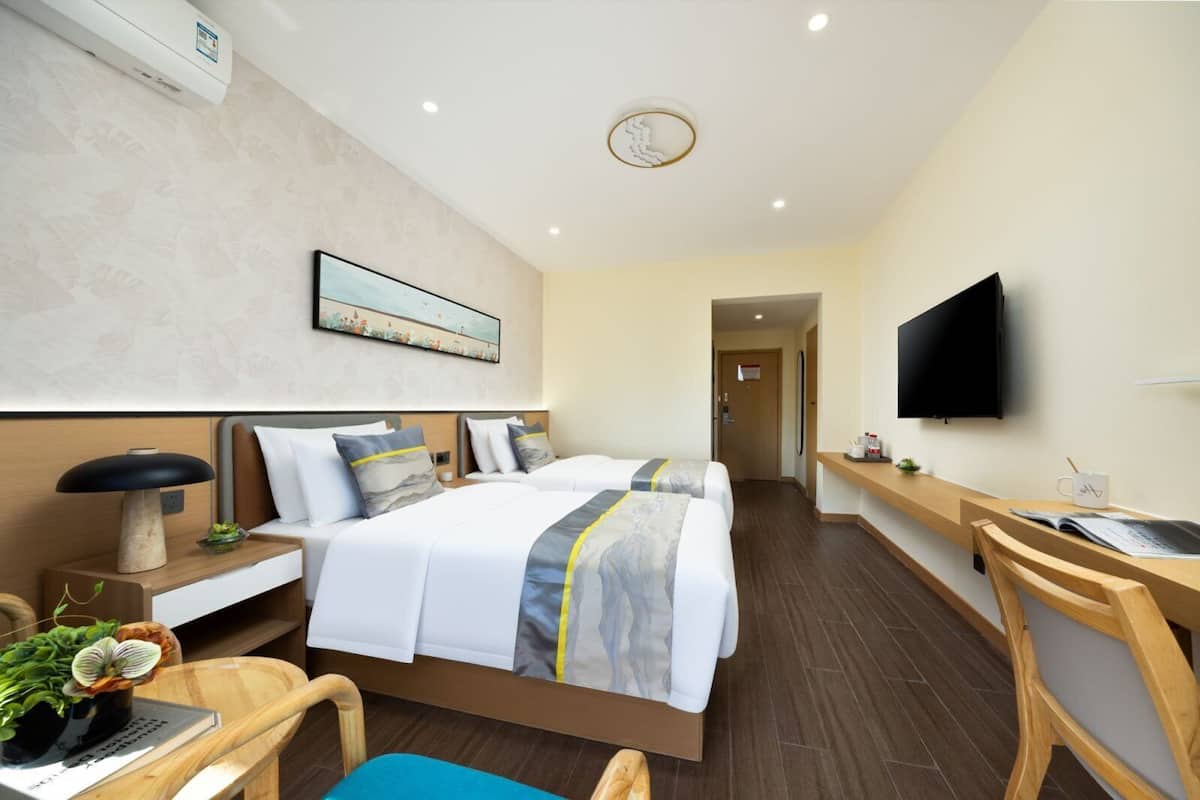 A hotel room with two beds, a desk, a chair, a TV, and a wall-mounted air conditioner.