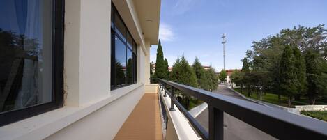 A balcony with a wooden floor, a metal railing, and a view of a street with trees and buildings.