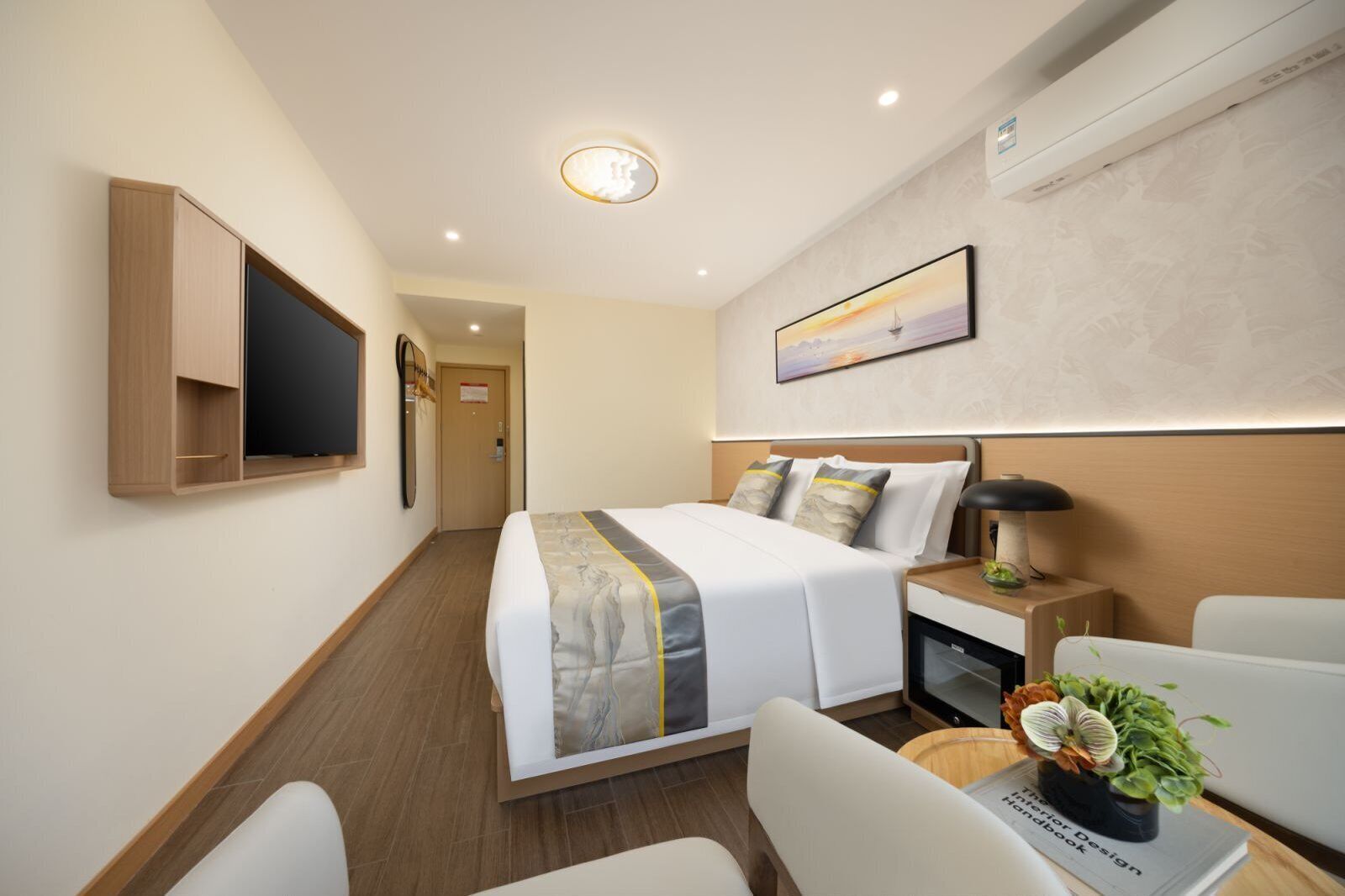 A hotel room with a bed, bedside table, lamp, chair, and a small potted plant.