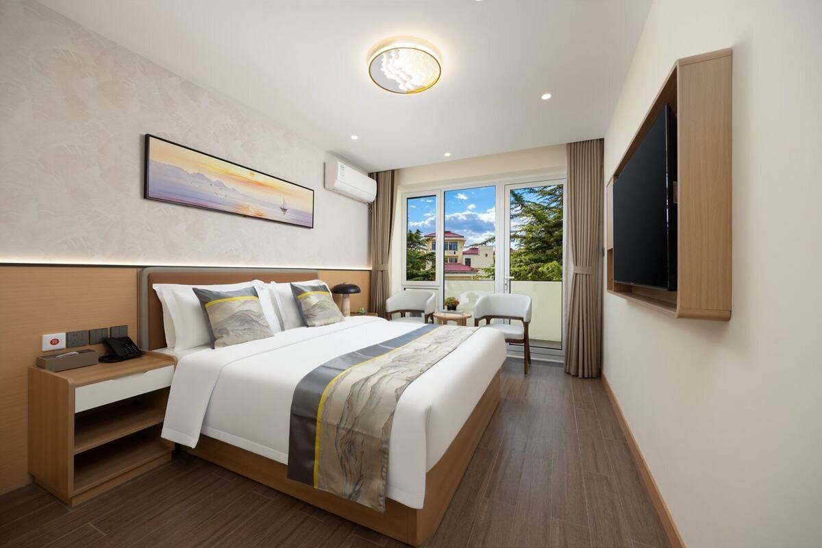 A hotel room with a large bed, bedside table, lamp, and a view of the outside.