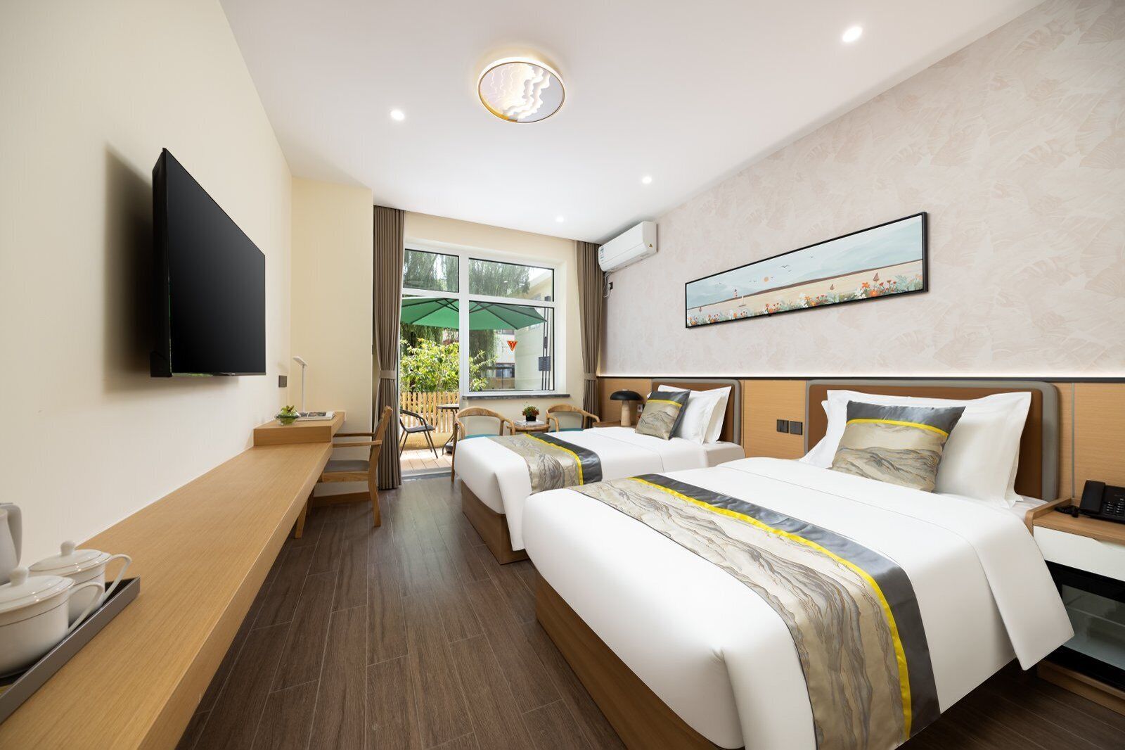 Quarto de hotel com duas camas, televisão, escrivaninha, cadeira e janela com vista para o verde.