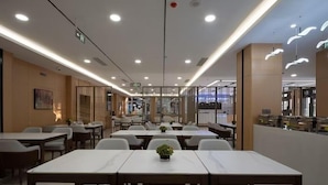 A modern dining area with multiple tables and chairs, a central plant, and a view of the cityscape through large windows. - Qinhuangdao Beidaihe Yijing Hotel (Qinhuangdao)