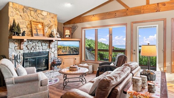 A spacious living room with a stone fireplace, leather sofas, a television, and large windows offering a view of the outdoors.