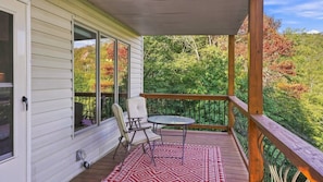 A covered porch with a table and chairs, a patterned rug, and a view of a forested area.