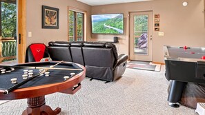 Ein Billardtisch mit roter Tasche, eine Couch, ein Fernseher und ein Kicker stehen in einem Raum mit Teppichboden und einer Tür zum Außenbereich.