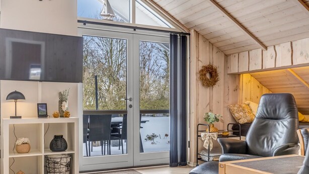Een modern interieur met een houten plafond, een zithoek met een leren fauteuil, een wit wandrek en een glazen deur die naar buiten leidt.