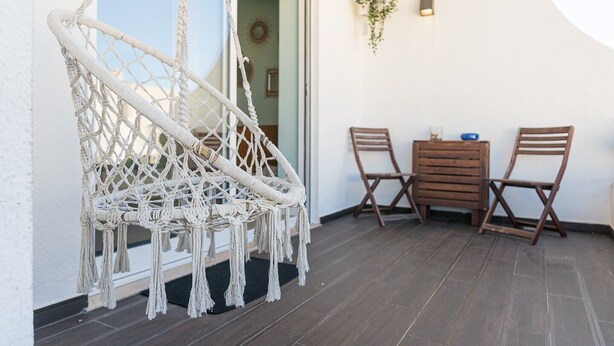 A macrame hammock chair, two wooden chairs, a wooden chest of drawers, and a small potted plant on a balcony with a dark wooden floor. - GuestReady - Relaxing escape near the beach (Moledo)