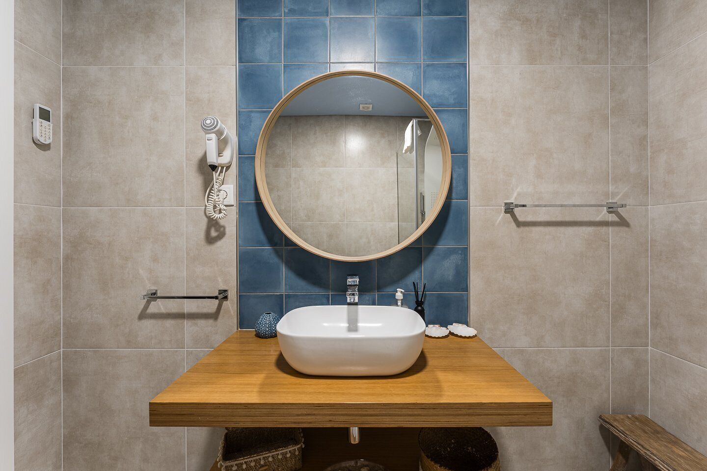 Een badkamer met een houten wastafelmeubel, een ronde spiegel, een witte wasbak en een blauwe betegelde muur.