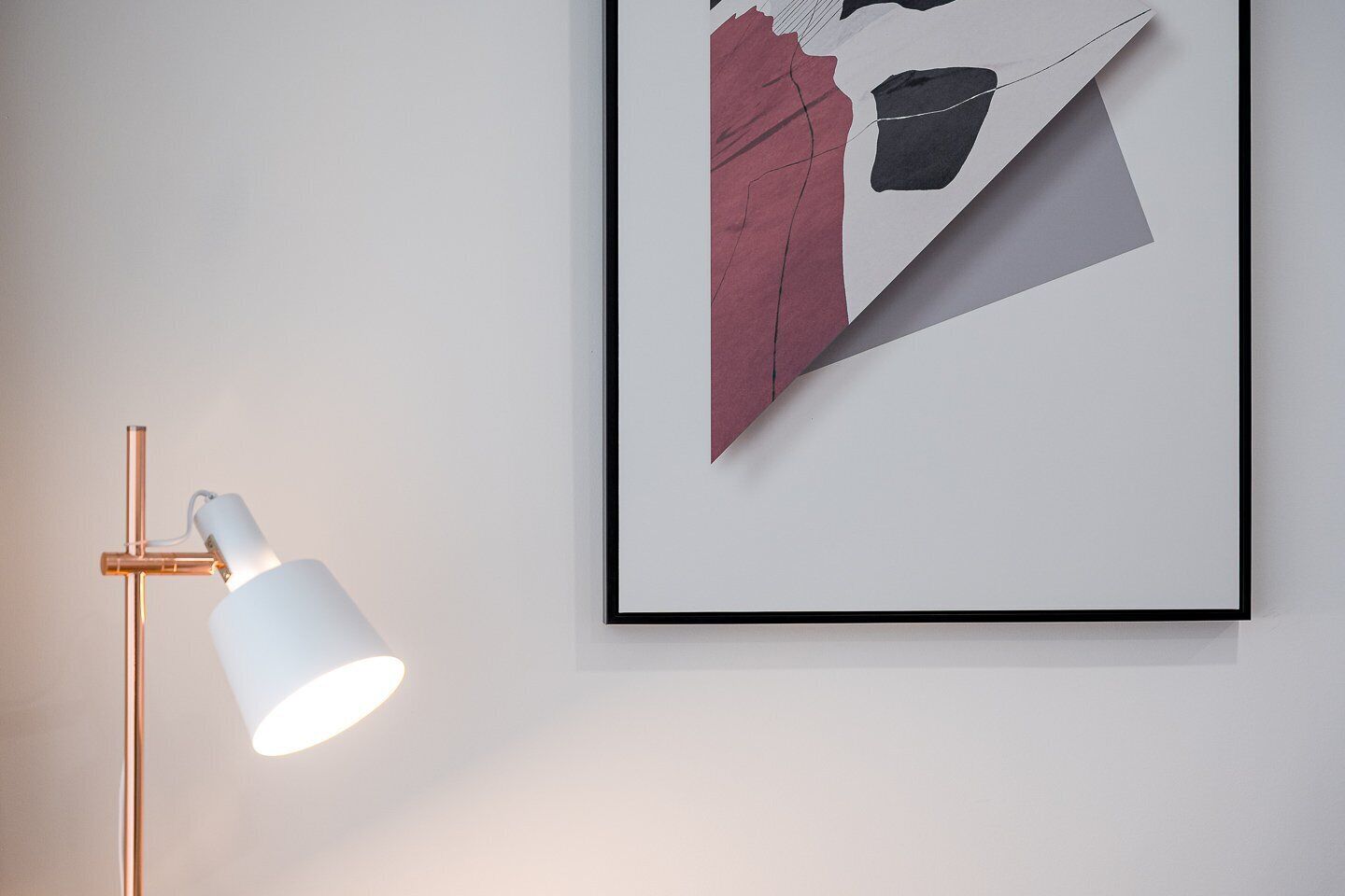 Een lijst met abstract kunstwerk aan een muur, een moderne vloerlamp en een effen muur.